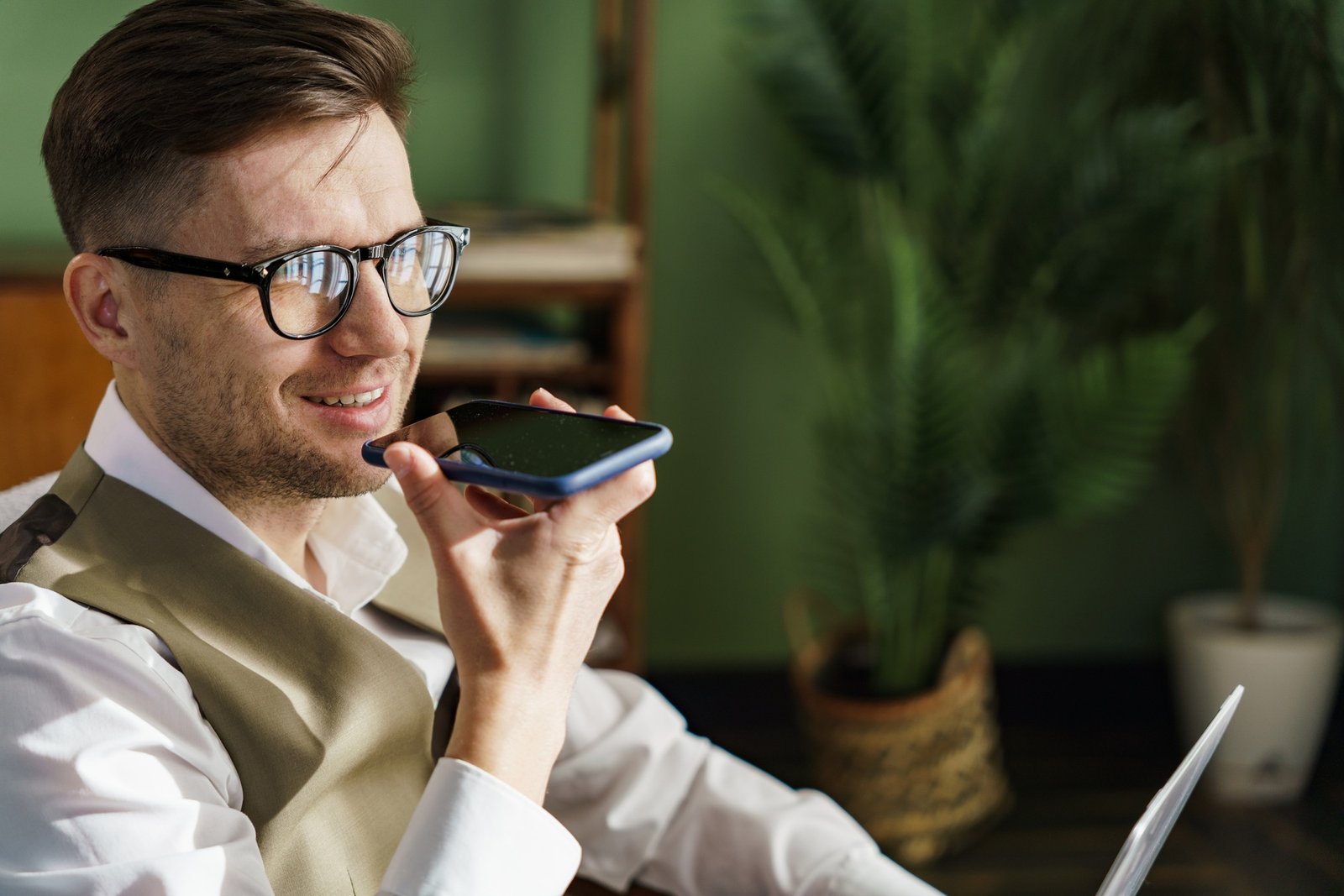 Businessman in smart vest using voice command on phone, indoor with plant backdrop.
