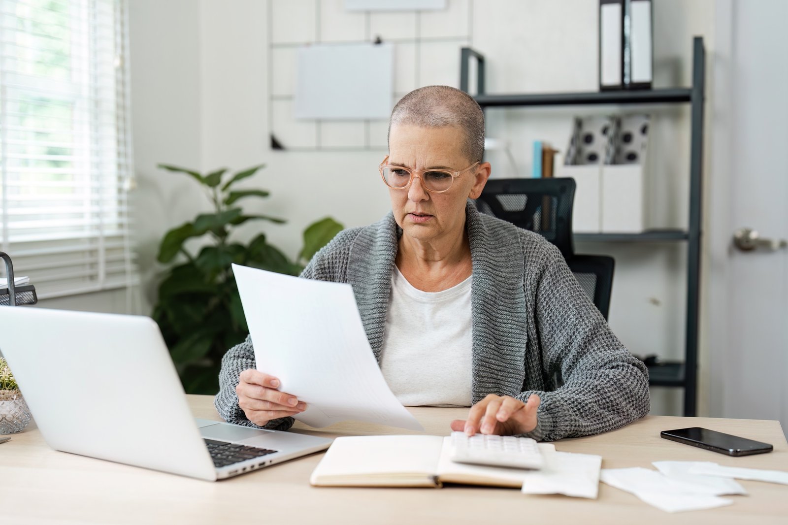Senior Woman Reviewing Financial Documents at Home Office Desk with Laptop and Calculator