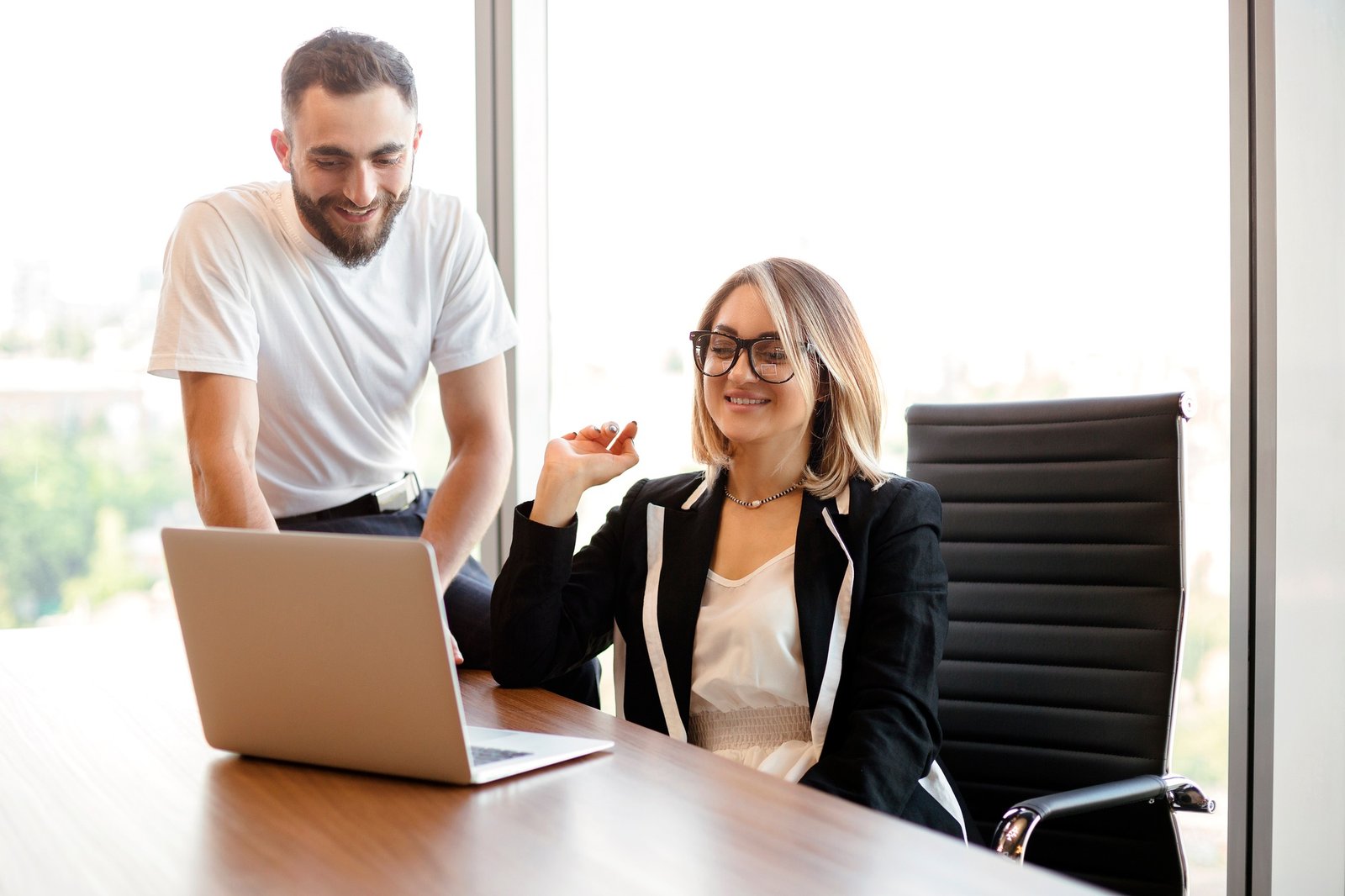 Positive businesswoman looking at laptop screen sitting at desk, training new male employee